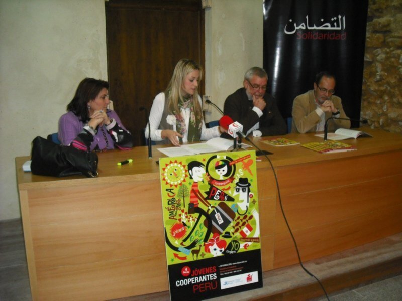 Una conferencia de prensa en una sala con una mesa de madera, una persona con un micrófono y una persona con una camiseta roja. La mesa tiene un cartel con el texto 'Jóvenes Cooperantes' y 'Solidaridad'.
