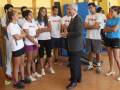 Un grupo de jóvenes vestidos con camisetas Wilson se preparan para un entrenamiento en una sala de deportes. Un hombre mayor, probablemente el entrenador, les da instrucciones mientras ellos miran atentos. La escena transmite un ambiente de concentración y preparación para una actividad física intensa.
