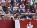 Un grupo de personas en una tribuna, algunos con bufandas y otros con gafas. Una persona está tomando una foto o grabando algo en el centro de la imagen. La estructura de la tribuna es roja con un logo blanco en el frente.