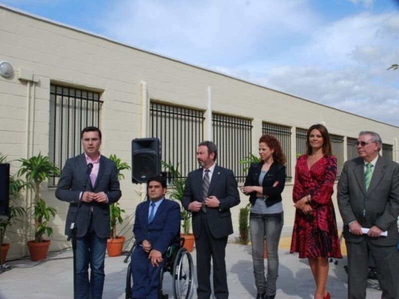 Persona en silla de ruedas junto a otros individuos en una presentación al aire libre frente a un edificio industrial.