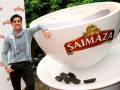 Un hombre posa junto a una gigante taza de café Saimaza, con granos de café al lado.