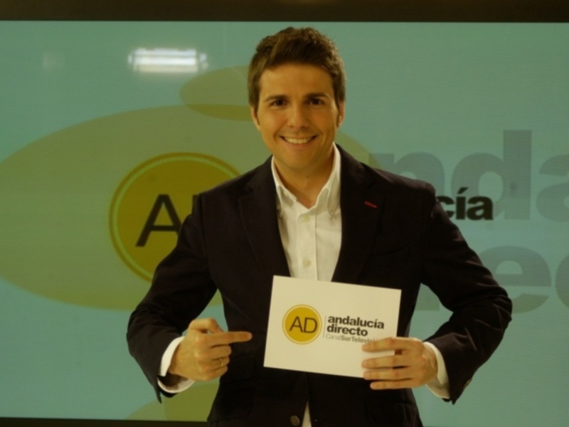 Un hombre en traje negro y camisa blanca sostiene un papel con el logotipo de Andalucía Directo en una presentación.
