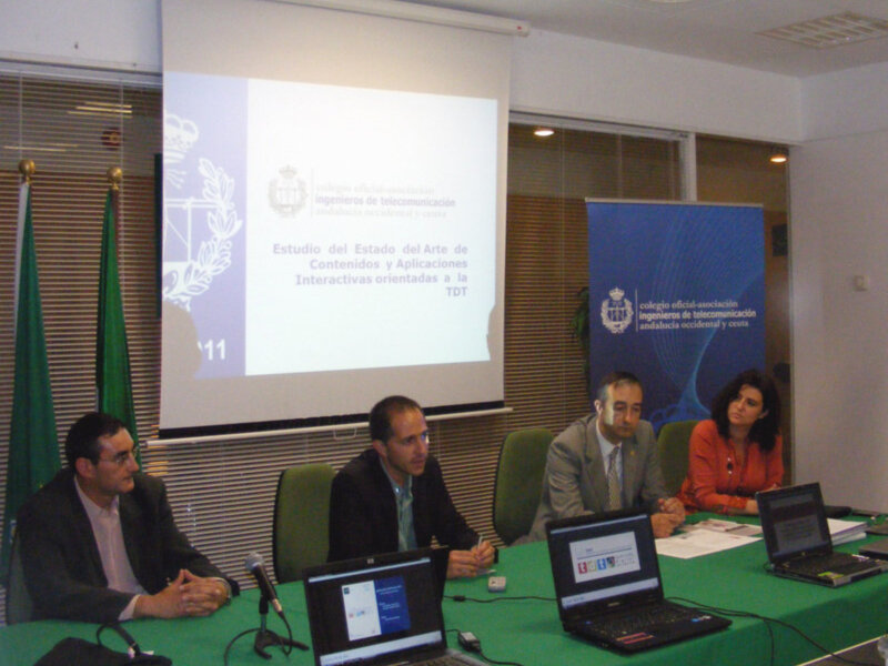 Presentación en sala de conferencias con tres personas sentadas en sillas frente a un proyector con un póster de la Asociación de Ingenieros de Telecomunicación en la pared.