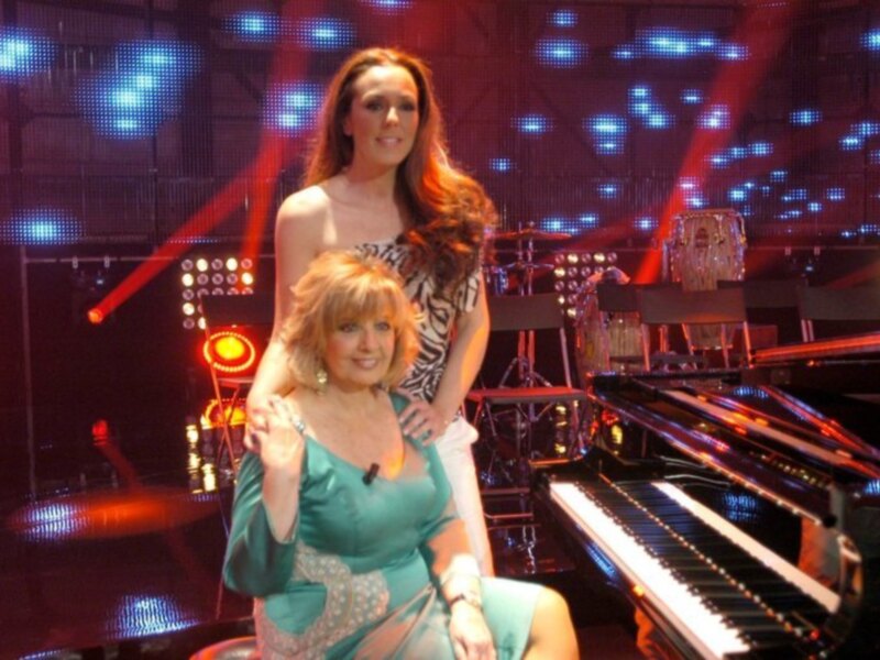 Dos mujeres posando en un escenario con un piano en el fondo, iluminado por luces azules y rojas.