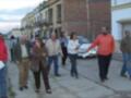 Una fila de personas caminan por una calle con edificios antiguos a ambos lados. Algunas personas llevan papeles o documentos en sus manos, mientras otras parecen estar conversando. El ambiente es urbano y parece ser un día soleado.