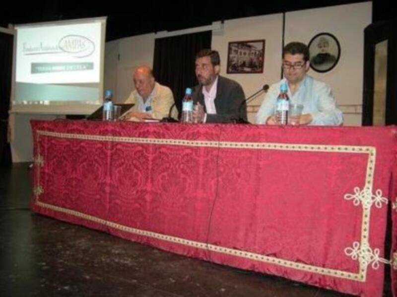 Tres hombres sentados en una mesa con una pantalla de presentación detrás, probablemente en una conferencia o reunión.
