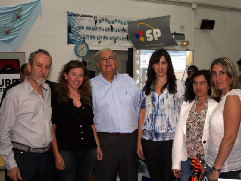 Grupo de personas en una oficina, con una señora con una camisa azul y blanca en el centro, y una señora en una camisa blanca a su derecha.