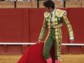 Un torero en traje tradicional de color verde y dorado, con guantes rosados, se prepara para la pelea. La arena está llena de espectadores que observan atentamente el evento.