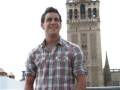 Un hombre joven con camisa a cuadros y pantalones vaqueros está sonriendo frente a la Giralda, una famosa torre en Sevilla.