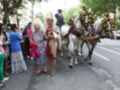 Una imagen festiva con personas vestidas de manera exagerada, incluyendo una anciana en un traje de novia y un hombre en sombrero. Dos caballos blancos con adornos florales se mueven por una calle, seguidos por un grupo de personas observando. La escena parece ser parte de una celebración o desfile festivo, con un ambiente alegre y colorido.