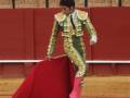 Un torero en traje verde y dorado con un capote rojo, preparándose para la lucha. La arena es de tierra y hay una cerca con espectadores detrás.