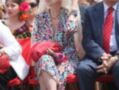 Una anciana con vestido floral y sombrero de plumas se sienta entre otros espectadores en un evento.