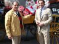 Dos hombres en trajes elegantes posan delante de una carreta con mujeres vestidas con trajes de flamenca en un entorno urbano.