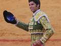 Un torero en su traje tradicional, con un sombrero azul y una saya amarilla, sonríe mientras sostiene su capote en la mano izquierda. El fondo es una arena de toros con suelo rojizo y líneas horizontales.