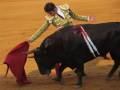 Un torero en traje tradicional se enfrenta a un gran toro negro en una plaza de toros. El torero sostiene una capa roja, mientras el toro se prepara para la lucha. La arena es de color amarillo brillante, típica de las plazas de toros españolas.