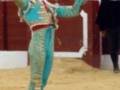Un torero en traje tradicional de color turquesa y rojo, con guantes y sombrero, levantando los puños en una plaza de toros.