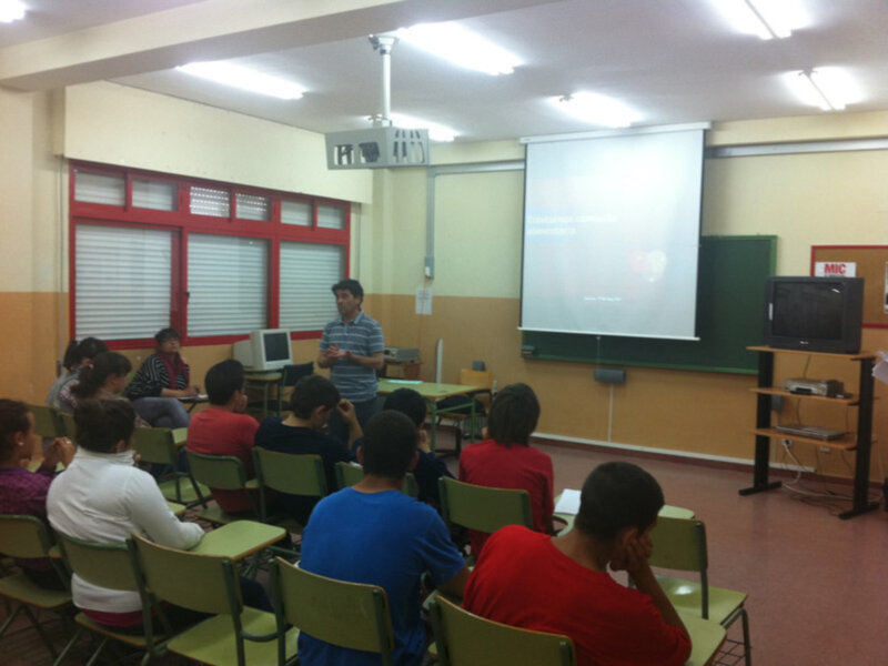 Un aula con estudiantes sentados en sillas verdes, un profesor en el frente y una pantalla proyectora en el piso.