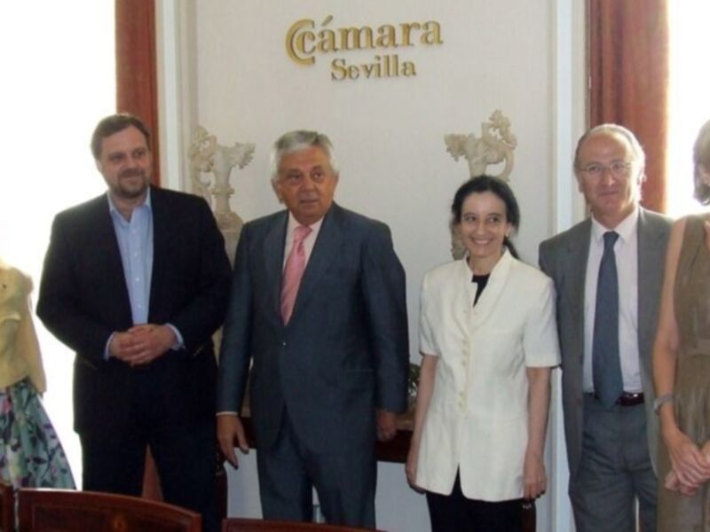 Una reunión formal en la Cámara de Sevilla con personas vestidas de negocios y una señora sonriente en el fondo.