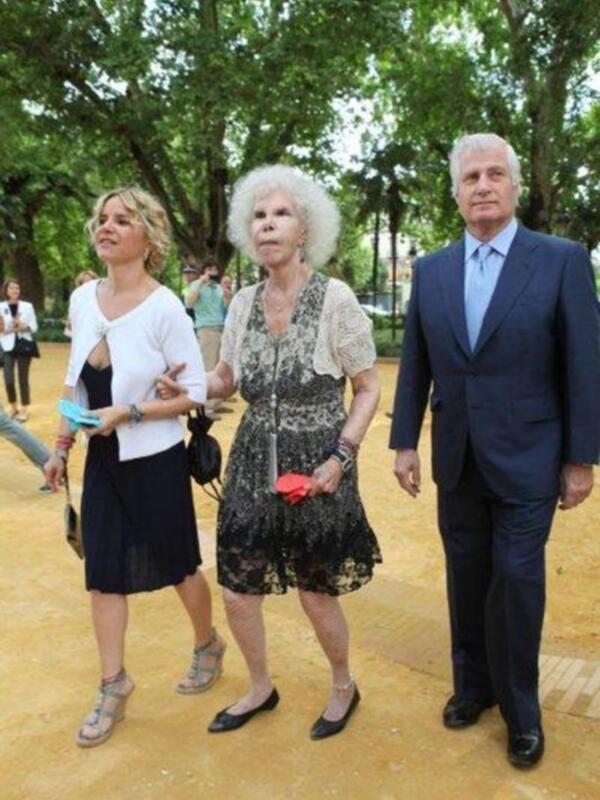 Una mujer con vestido negro y una mujer mayor con vestido de encaje y sombrero blanco caminan juntas en un parque con árboles verdes y un hombre en traje azul a su lado.
