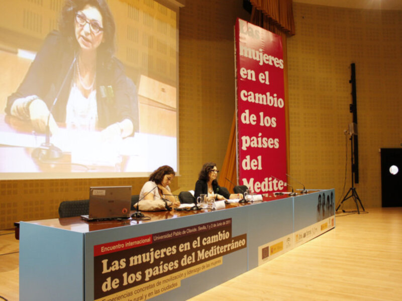 Dos personas sentadas en una mesa con un banner que dice 'Las mujeres en el cambio en los países del Mediterráneo' en el fondo.