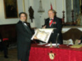 Dos hombres en trajes formales sostienen un diploma, frente a una mesa con mantel rojo y decoración. En el fondo, se observan cuadros y una estatua.