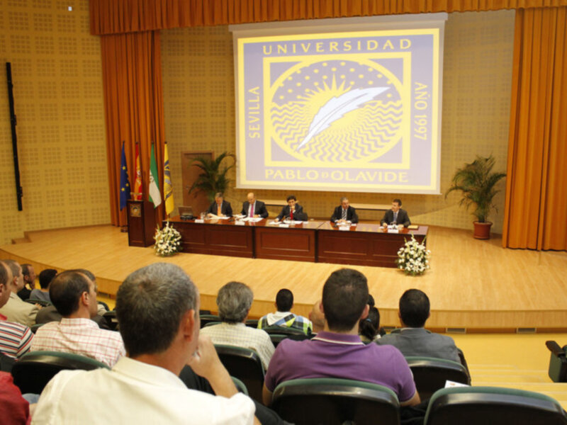 Conferencia en la Universidad Pablo de Olavide, Sevilla, con panelistas en el escenario y público asistente.