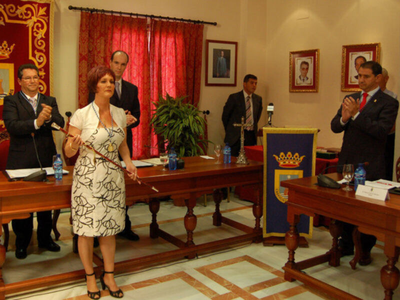 Mujer hablando en un acto oficial en un salón con cortinas rojas y retratos enmarcados en la pared.