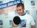 Un hombre con una camiseta de "Brugal" sostiene un micrófono en una presentación. El fondo tiene el texto "CASTING BRUGAL" y un lema sobre la participación en reportajes.