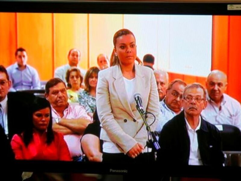 Una mujer en una conferencia de prensa, con un público detrás de ella, en un entorno formal.