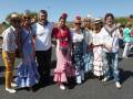 Un grupo de personas vestidas con trajes típicos de la Feria de Abril, posando en una carretera. La imagen muestra a hombres y mujeres con trajes de flamenca, sombreros y accesorios típicos. La escena parece ser en un día soleado, con cielo azul y algunos árboles en el fondo.