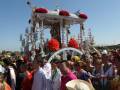 Una procesión religiosa con gente vestida de colores y sombreros, un carro adornado con flores rojas en el centro.