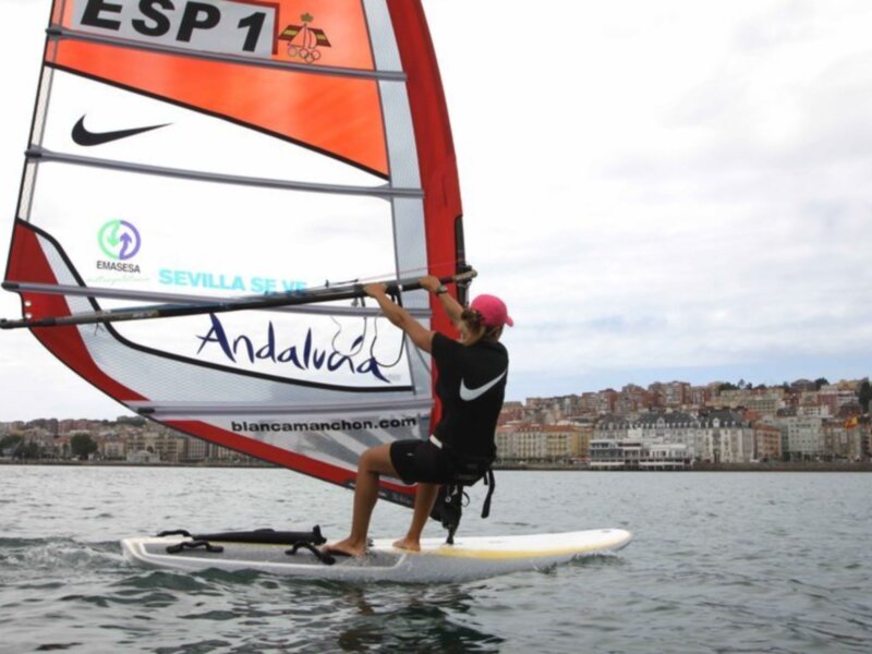 Un windsurfer en acción con el viento en la vela, en el mar frente a una ciudad costera.