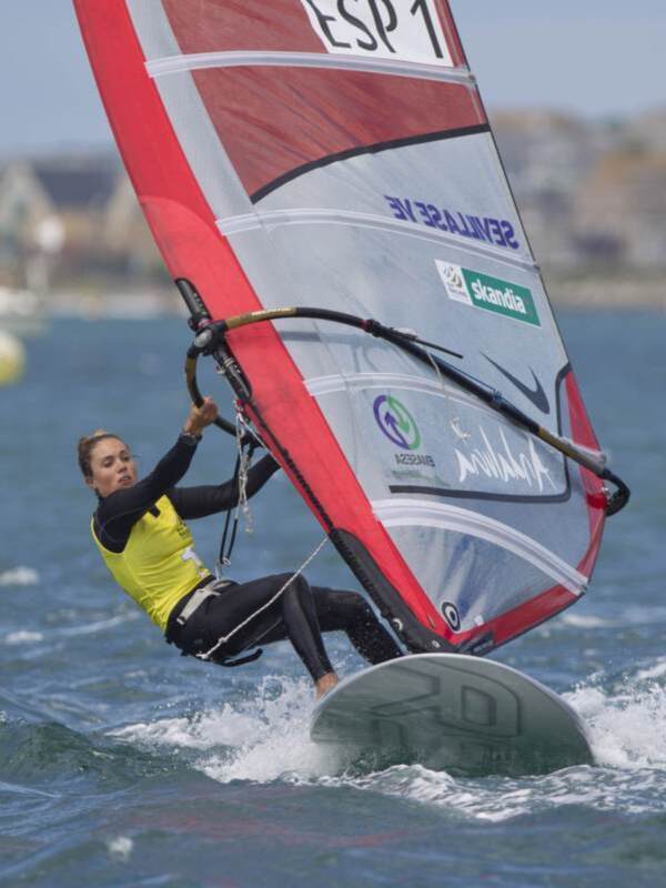 Una windsurfera en acción, con el viento fuerte y el mar azul. La imagen muestra la intensidad y la belleza del windsurfing en un día soleado.