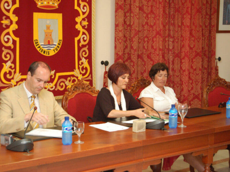 Tres personas sentadas en una mesa con portátiles, botellas de agua y vasos de cristal. La pared detrás de ellos tiene un escudo heráldico y cortinas rojas. El ambiente parece ser formal y de reunión.