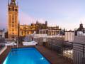 Una piscina con una vista panorámica de la Giralda, una torre icónica en el horizonte. La piscina está rodeada por un balcón con sillas blancas y una vista de la ciudad. El cielo es claro y el sol está a punto de ponerse, creando un ambiente tranquilo.