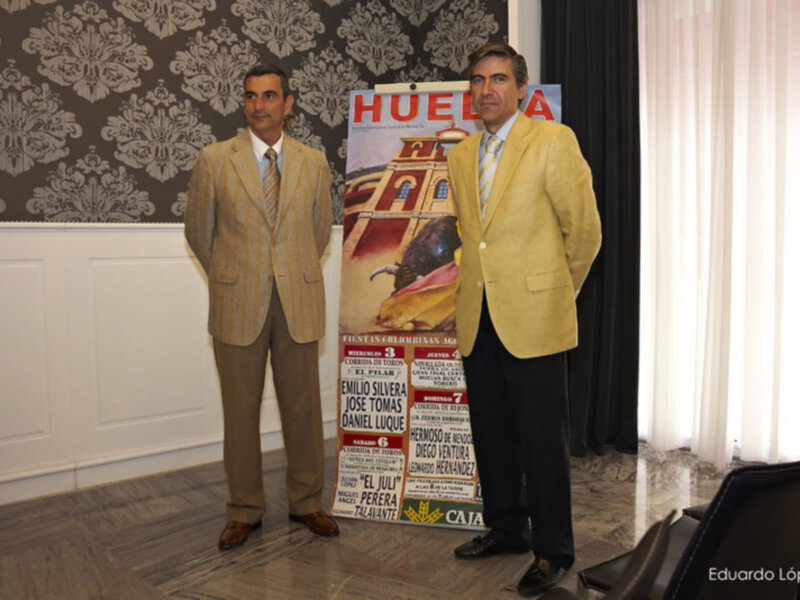 Dos hombres en traje posan junto a un cartel de toros en un evento formal.