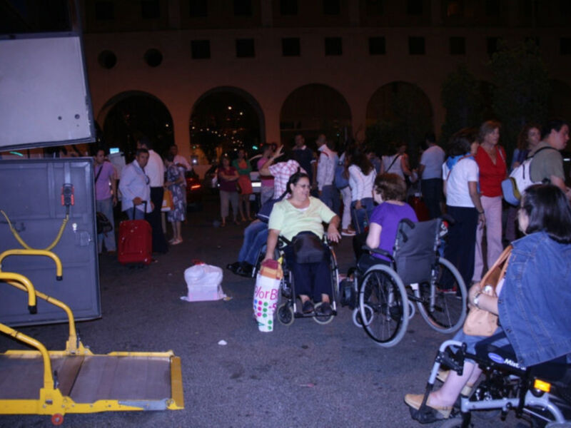 Una escena nocturna con personas en sillas de ruedas y bolsas de compras, posiblemente en un evento o manifestación.