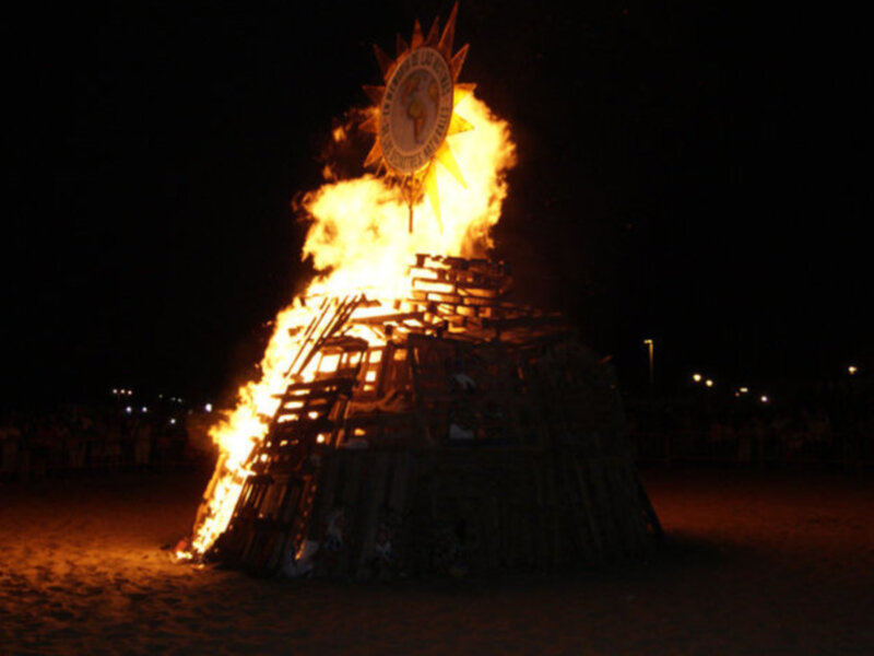Fuego ardiente en una estructura de madera en una playa nocturna, rodeada de espectadores.