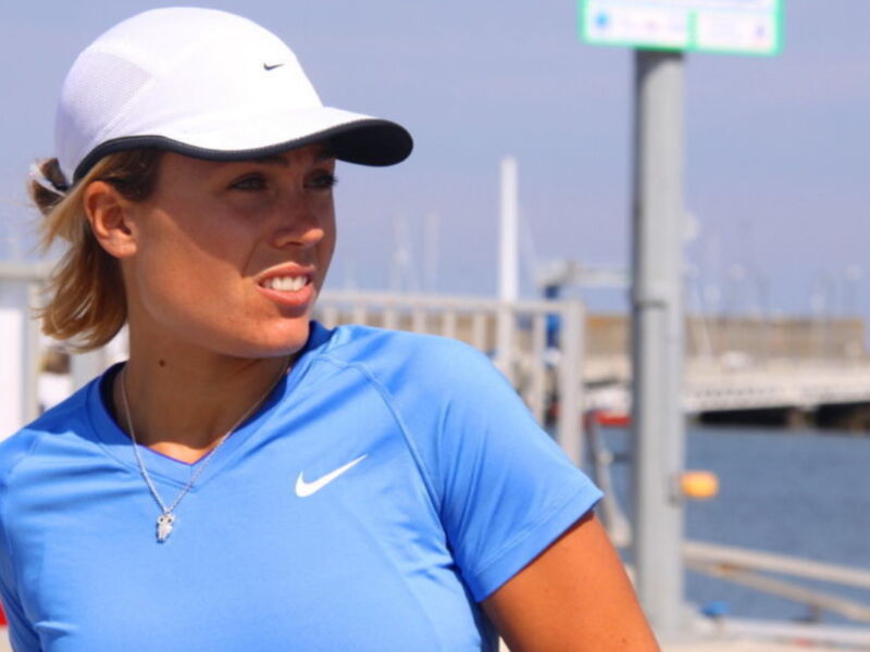 Una mujer con cabello recogido en una coleta, vistiendo una camiseta azul y una gorra blanca con un logotipo de Nike, posando frente a un mar tranquilo con un puente en el fondo.