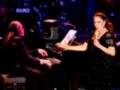 Una mujer cantante con micrófono en mano, frente a un pianista que toca el piano. La imagen muestra una escena de concierto con luces y un público en segundo plano.