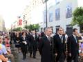 Desfile de oficiales en uniforme frente a un edificio con carteles de publicidad en la calle. El ambiente es formal y ceremonial, con personas observando desde el lado.