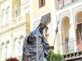Estatua de un santo en procesión, rodeada por flores y antecedentes históricos.