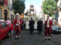 Imagen muestra una procesión religiosa con hombres vestidos en trajes rojos y blancos, sosteniendo bastones de procesión. En el fondo hay una carroza con flores y un altar en la parte superior. La escena se desarrolla en una calle con edificios y coches a ambos lados.