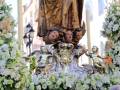 Una estatua religiosa rodeada de flores blancas y verdes, posiblemente durante una procesión o celebración. La estatua parece ser de un personaje importante, posiblemente una virgen o santo, y está decorada con un escudo de armas en el frente. La atmósfera es festiva y la imagen transmite una sensación de tradición y devoción.