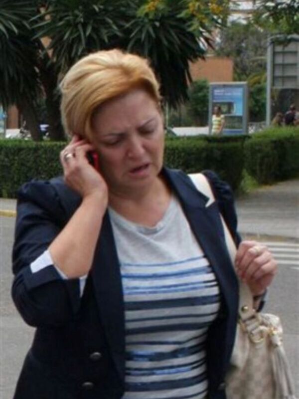 Mujer hablando por teléfono móvil mientras camina por una calle con arbustos y edificios al fondo.