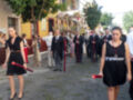 Procesión religiosa con personas portando palos rojos, vestidas de negro, en una calle de pueblo con edificios y árboles al fondo.