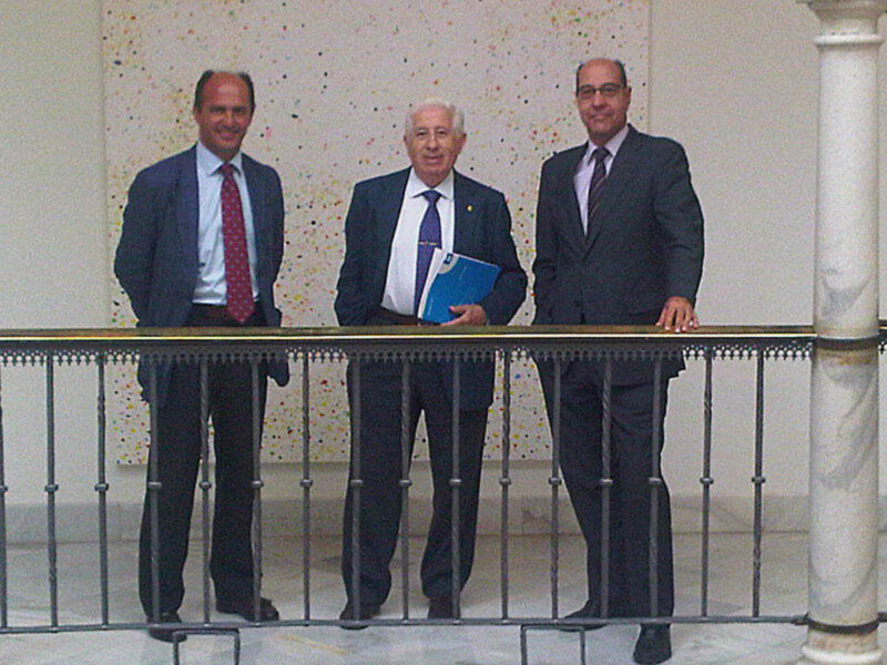 Tres hombres en traje posan juntos en un balcón, uno sostiene un libro azul.