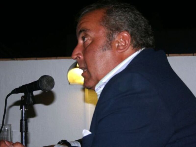 Hombre hablando en un evento formal con un microfono en frente de él y una copa de vidrio en la mano. El fondo es oscuro y la iluminación es suave, enfocándose en el hombre y el microfono.