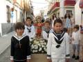 Niños vestidos de marinero participan en una procesión religiosa, rodeados de flores y banderas.
