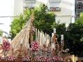 Una procesión religiosa con una figura vestida de blanco y dorado, posiblemente una imagen de la Virgen María, acompañada por velas y flores en un ambiente urbano con edificios y árboles al fondo.